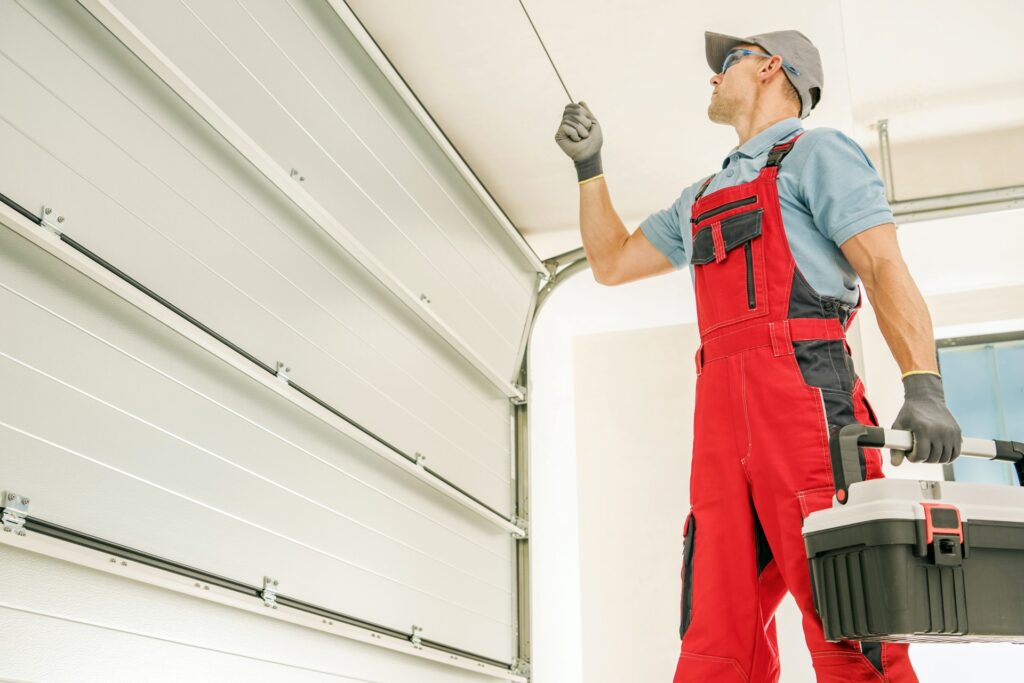 A technician in red bib overalls and a gray cap pulls the emergency release cord of a white sectional garage door while carrying a black tool case.