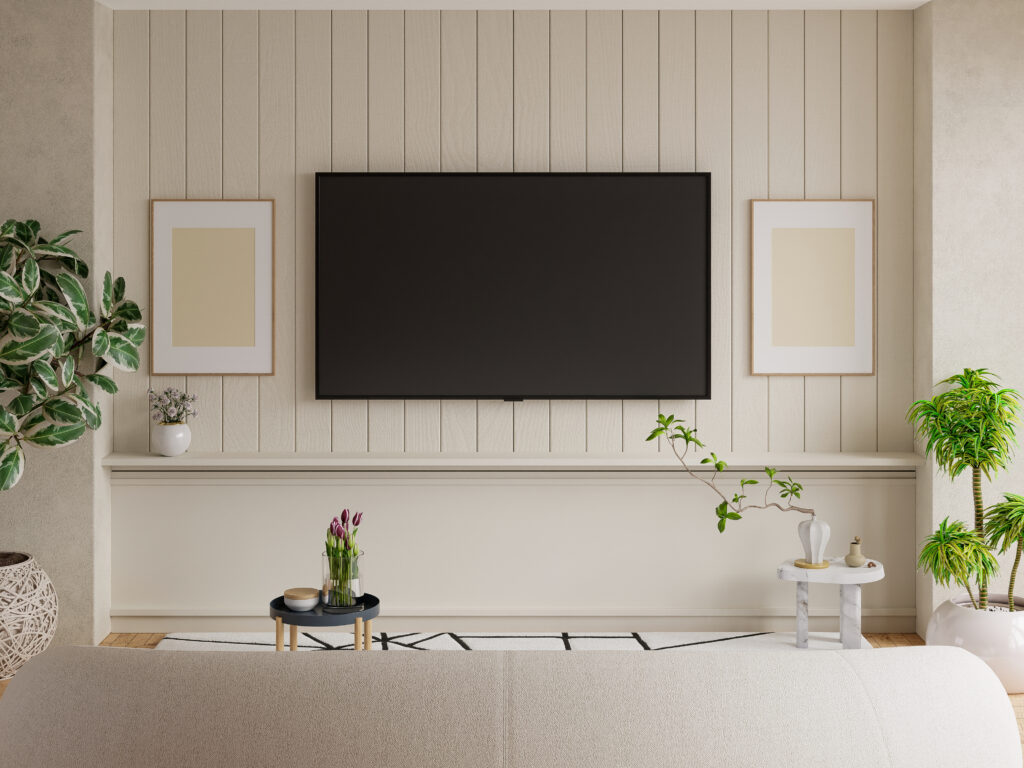 A minimalist living room interior featuring a large wall-mounted flat screen TV on a beige vertical panel wall, flanked by two framed art prints, with houseplants, a marble side table, tulips on a navy coffee table, and a cream sofa on a patterned rug.